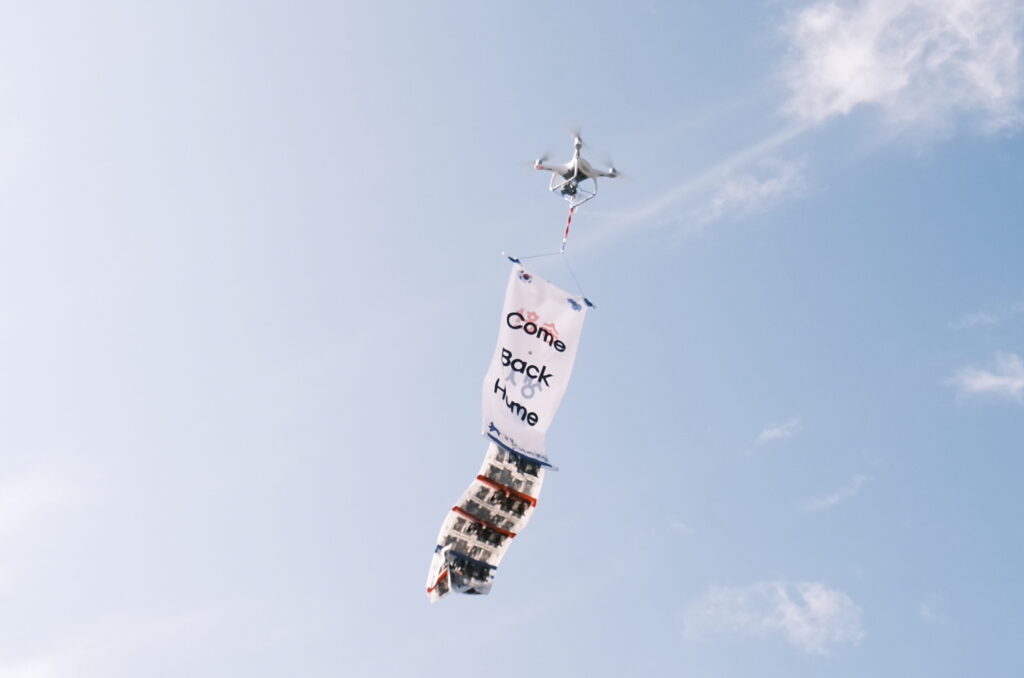 A drone floats in a blue sky with a banner beneath it with the phrase in English "Come Back Home."
