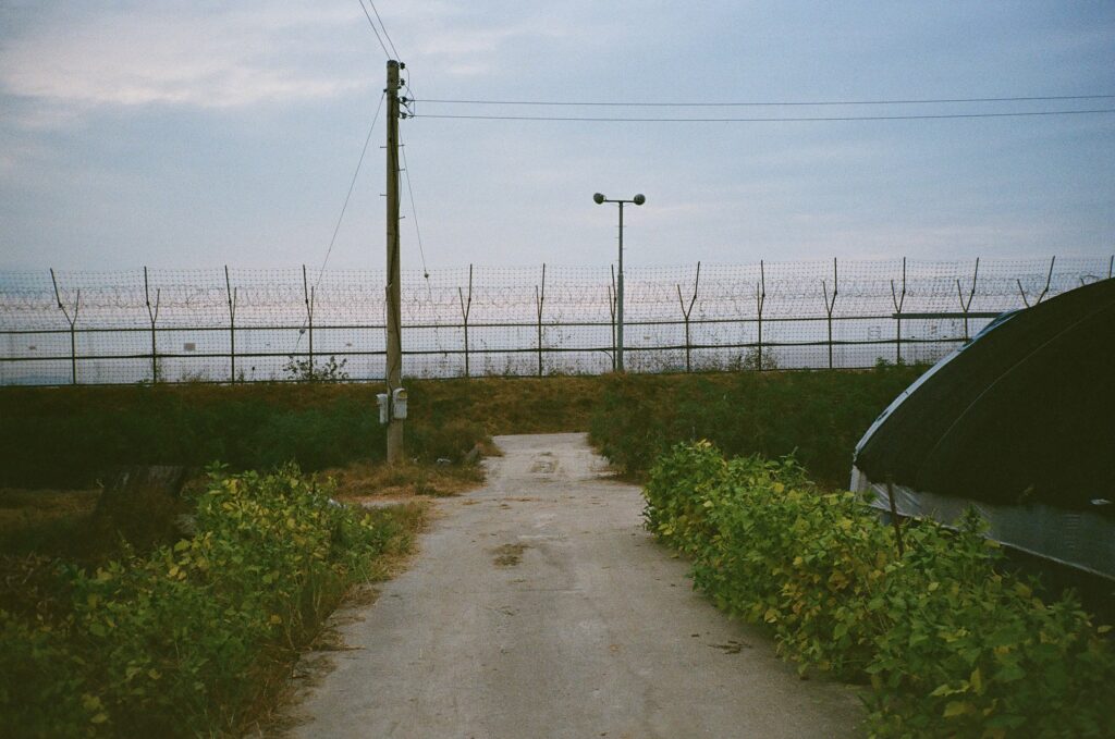 A road ends at a fence, which also includes a pole with speakers on top of it.