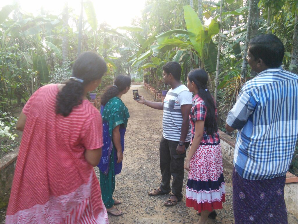 On a dirt and gravel trail amid verdant palm leaves, a man in a white and blue striped T-shirt holds up a smartphone as other members of his family gather around.