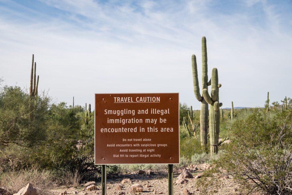Amid rocky ground, low shrubs, and spiky cacti beneath a pale blue and white sky, a brown sign reads: “Travel Caution. Smuggling and illegal immigration may be encountered in this area.”