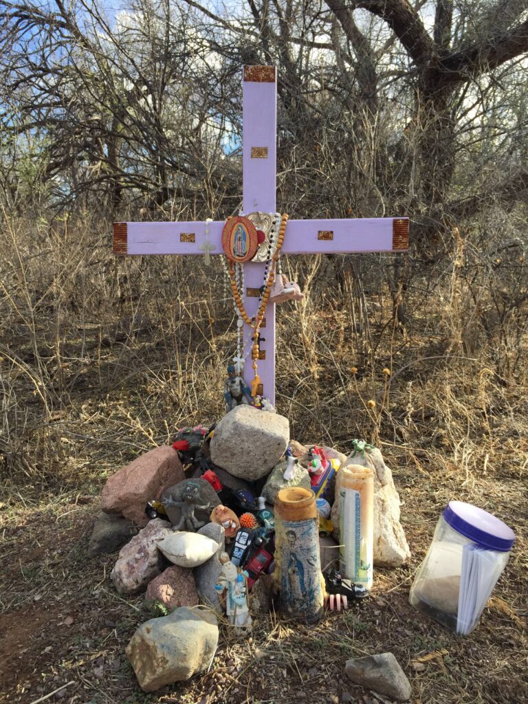 A white cross standing near trees and low brush is surrounded by rocks, candles, and other memorabilia.