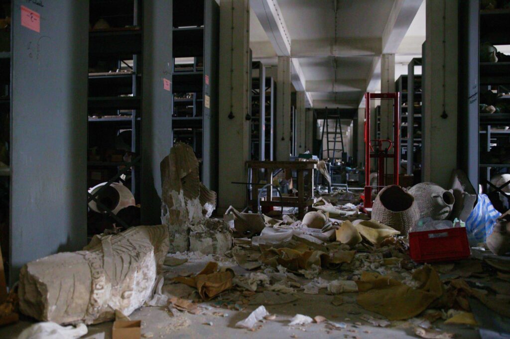 Smashed clay pots and other objects lie scattered in a long, poorly lit hallway that contains rows of shelves.