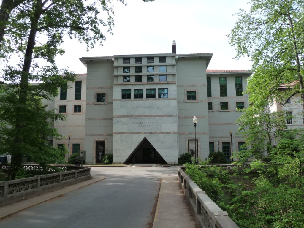 A building with a light-beige facade and triangular entryway stands at the end of a paved path lined with tall, thin trees and low shrubs.
