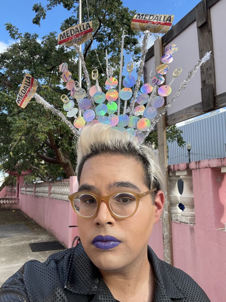 Una persona con gafas de montura de plástico beige, pintalabios morado intenso, pelo corto decolorado y una tiara casera mira a la cámara con un ligero aire de rebeldía.