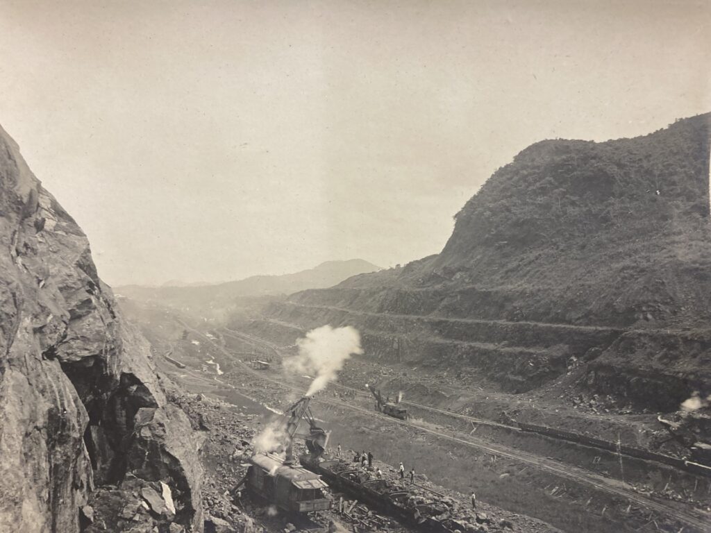 An old sepia-toned photograph shows steam-powered industrial machinery working in a valley between two large rocky uprisings.