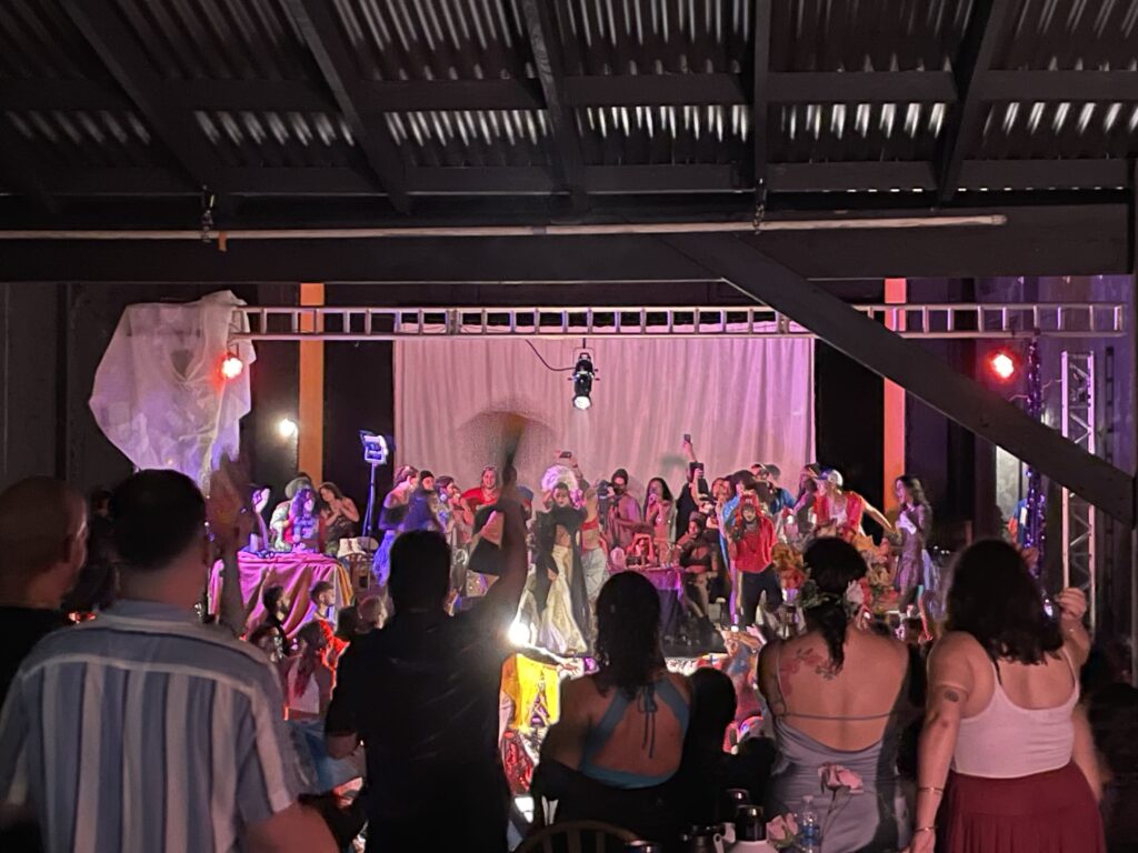 Una multitud de espectadores observa a un numeroso grupo de personas vestidas con ropa de colores vivos sobre un escenario iluminado por focos y con un techo negro.