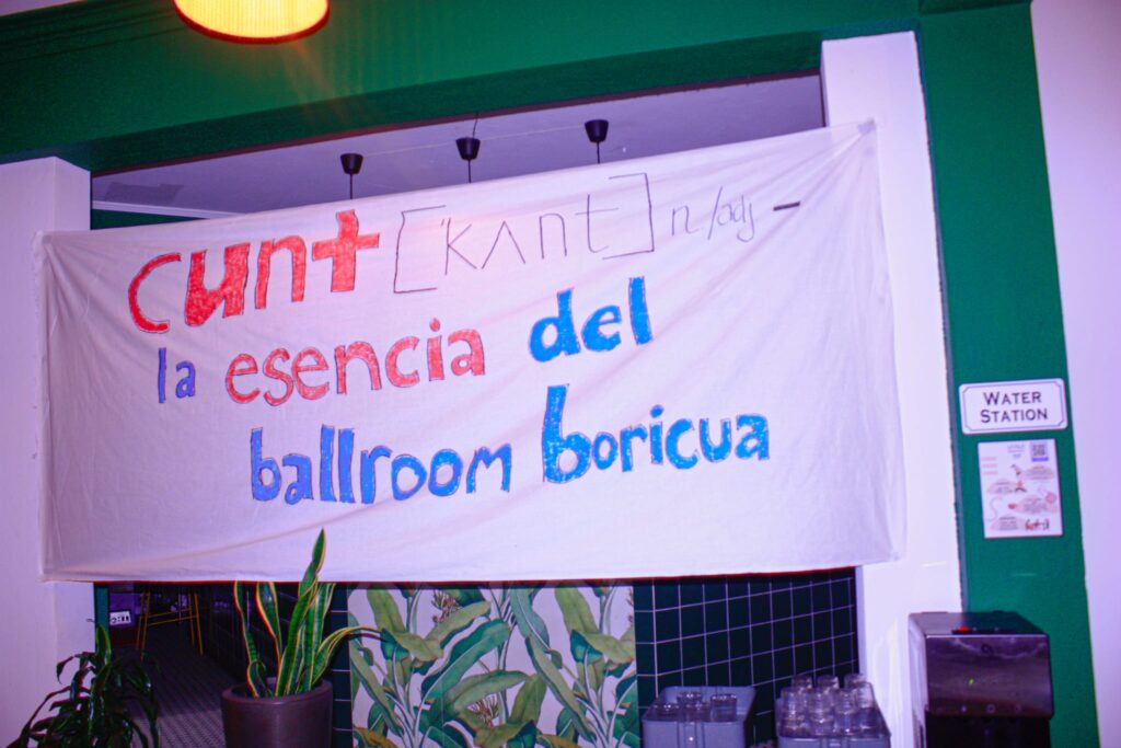 Un cartel casero pintado en una sábana blanca con letras rojas, naranjas y azules dice: "Cunt—la esencia del ballroom boricua".