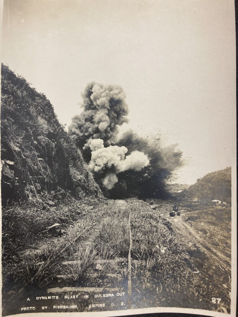 In a sepia-toned photograph, train tracks along a grassy area beside a hill approach an area where a demolition explosion throws large clouds of debris into the air.
