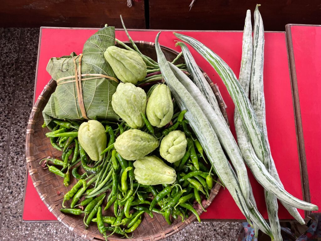 A wicker basket filled with green peppers and other green vegetables sits on a red surface. Beside the bowls are long green stalks.