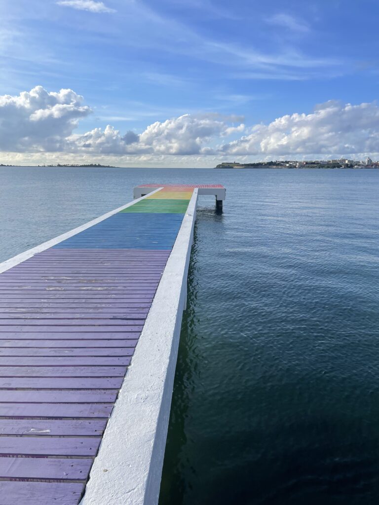 Un largo muelle pintado con los colores del arcoíris se adentra en unas tranquilas aguas. Islas y grandes nubes blancas y esponjosas salpican el horizonte.