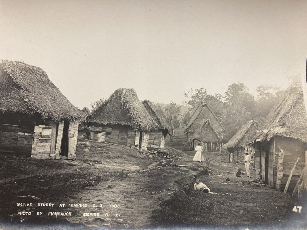 A sepia toned photograph shows people amidst wooden thatched-roof structures.