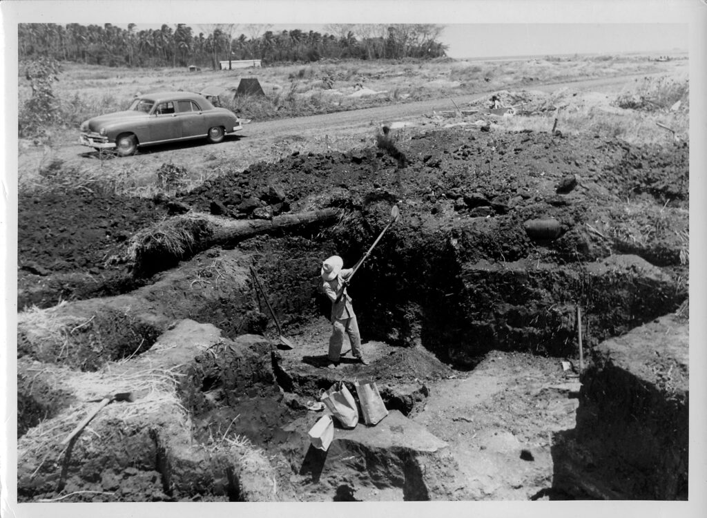 In a black-and-white photograph a person in a broad-brimmed hat shovels dirt while standing in the middle of an excavation site. An automobile is parked in the background.