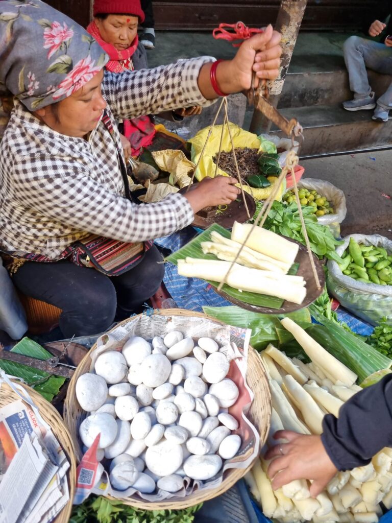 Sitting next to a table, a person with a flowered scarf tied around their head holds up a scale full of food. A hand reaches out to touch piles and containers of other goods that sit nearby.