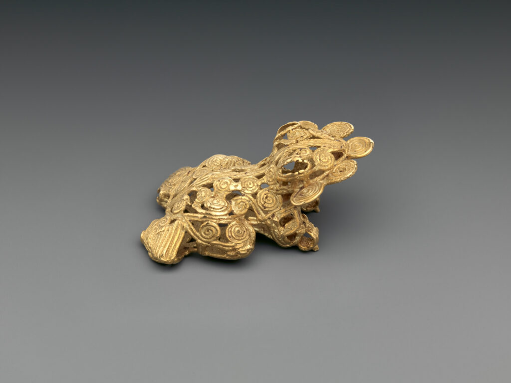 A piece of intricately constructed gold jewelry shaped like a frog sits on a gray surface.