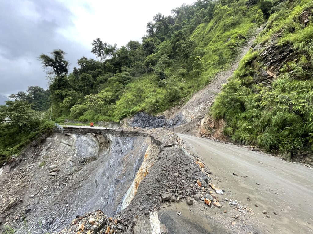 Behind a heavily damaged ribbon of gray road covered in piles of debris, a steep hill rises, covered in thick green growth.