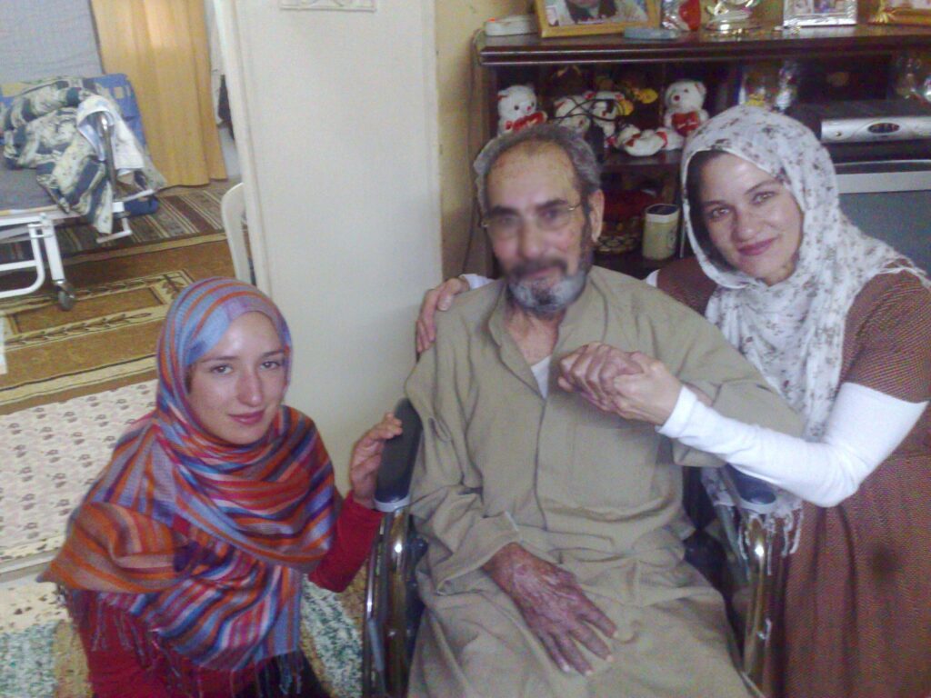 A man in a tan thobe sits in a wheelchair next to a woman in a brightly colored hijab on one side and another woman in a brown and white hijab and dress on the other side who holds the man’s hand. A medical bed is in the backdrop.