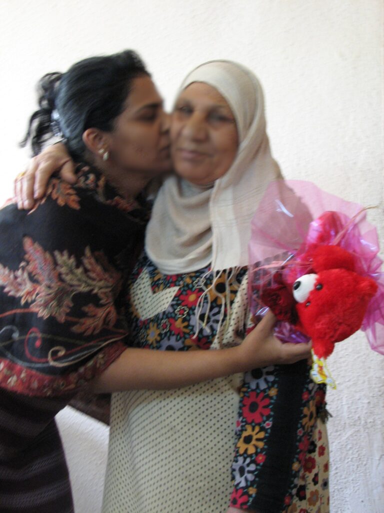 A woman on the left with black hair and a black abaya holds a red teddy bear while hugging a woman on the right who wears a white hijab and a white, dotted abaya.