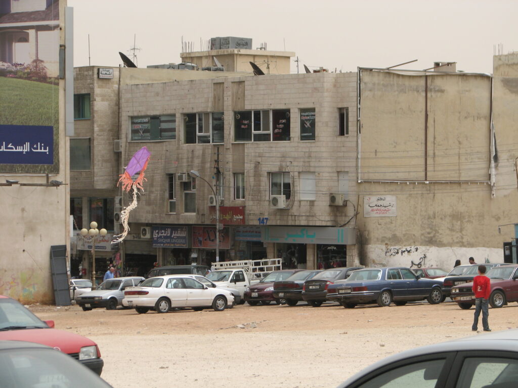 A kite with a white tail hovers between tan brick buildings and over rows of parked cars. A young person in a red shirt stands on the right side of the photo.