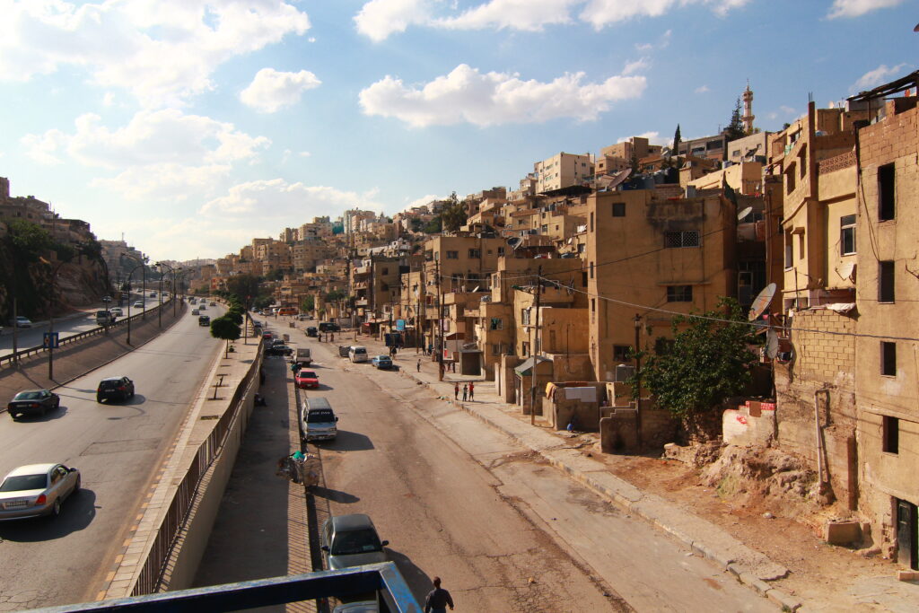 A view of a highway on the left features crowded, earth-colored brick buildings on the right that abut a street lined with parked cars.