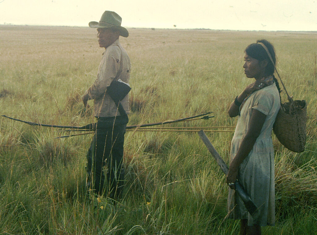 A man and a woman stand in a savanna carrying foraging gear. The man holds a bow and arrows, and tucked under his arm is a stork hat worn as camouflage to hunt deer. The woman carries a digging stick and woven bag on her back with the strap slung across her head.