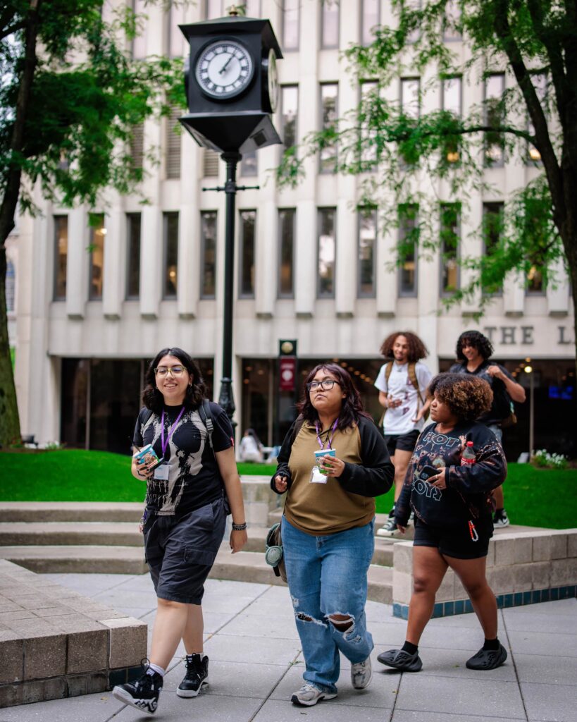 Five young people stroll in front of a gray concrete building with numerous narrow rectangular windows. In the background are leafy trees and a clock affixed to a pole.