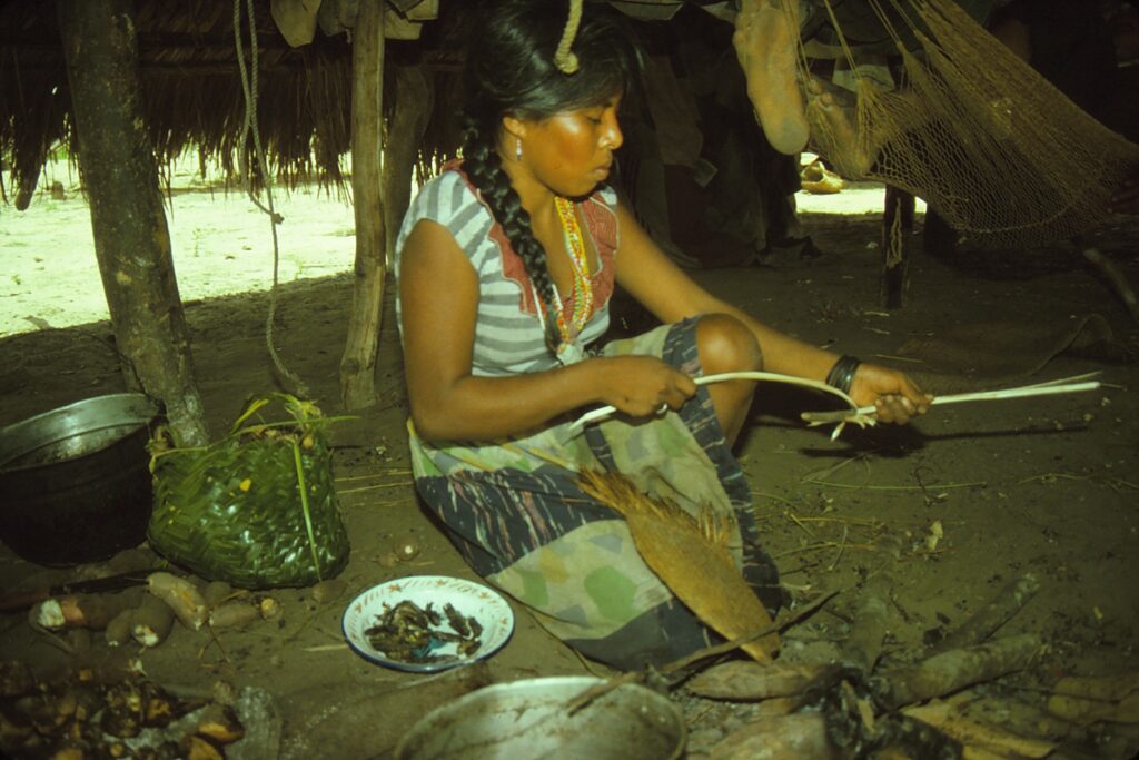 A person with a long black braid sits on a dirt floor in a thatch hut stripping bark. A palm fiber hammock is behind her, a fire fan lies in her lap, and a woven basket rests beside her.