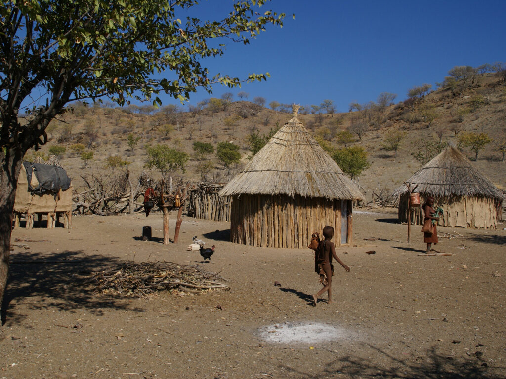 People stroll through a small village of wooden huts. Except for a tree, the landscape is beige and rocky, with trees growing sparsely on the hills that rise behind the village.