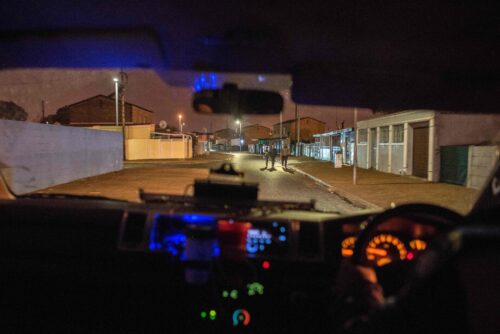 Trois personnes visibles à travers le pare-brise d'une voiture dont le tableau de bord est éclairé par des lumières orange, rouges, vertes et bleues marchent dans une rue la nuit.