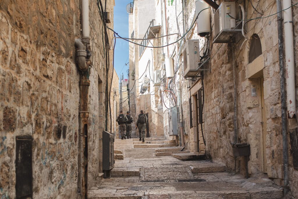Três pessoas vestidas com uniformes cinza e pretos patrulham uma rua estreita que passa entre prédios de pedra bege.