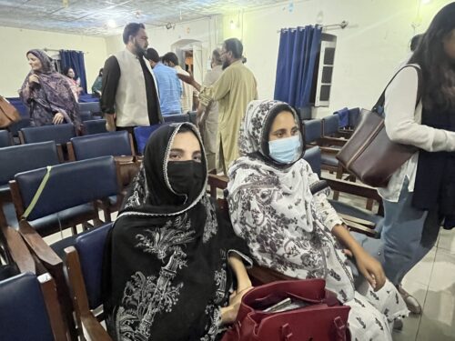 Two women wearing headscarves, face masks, and black and white dresses called “shalwar kameez” sit beside each other in a room with rows of blue-upholstered chairs as a handful of other people mill about.