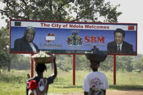 Two people carrying baskets on their heads look up at a billboard showing the faces of two men.