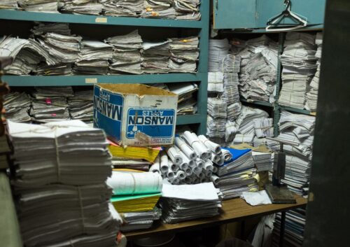 In a small room, stacks and rolls of papers fill teal-colored shelves and cover a table.