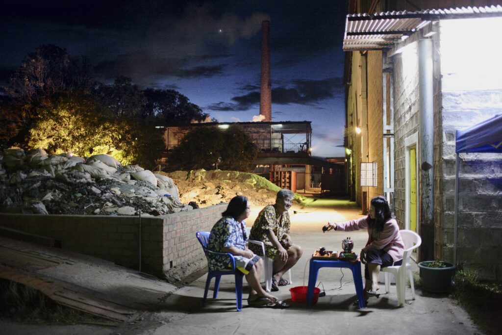 Iluminadas por luzes elétricas, três pessoas sentam-se em torno de uma pequena mesa ao ar livre, enquanto nuvens flutuam no céu noturno acima delas.