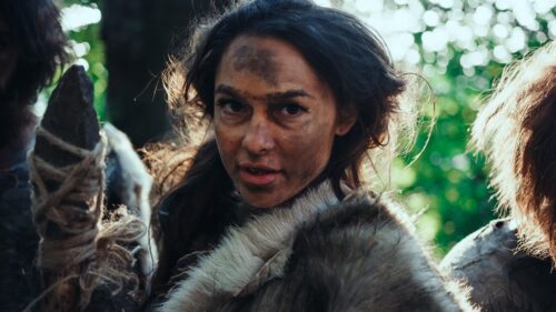 A person with long brown hair and a smudge of dirt on her forehead carries a stone weapon and wears a fur around her shoulders and neck.