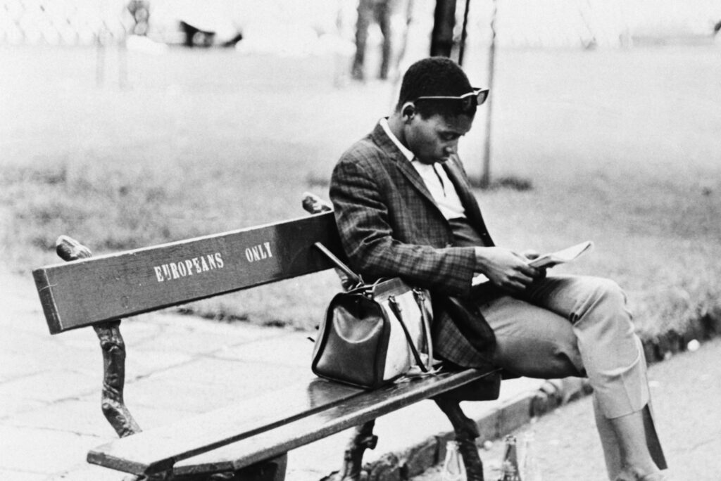 A black-and-white photograph shows a Black person sitting on a park bench, legs crossed, reading. On the bench, stenciled letters say: “Europeans Only.”