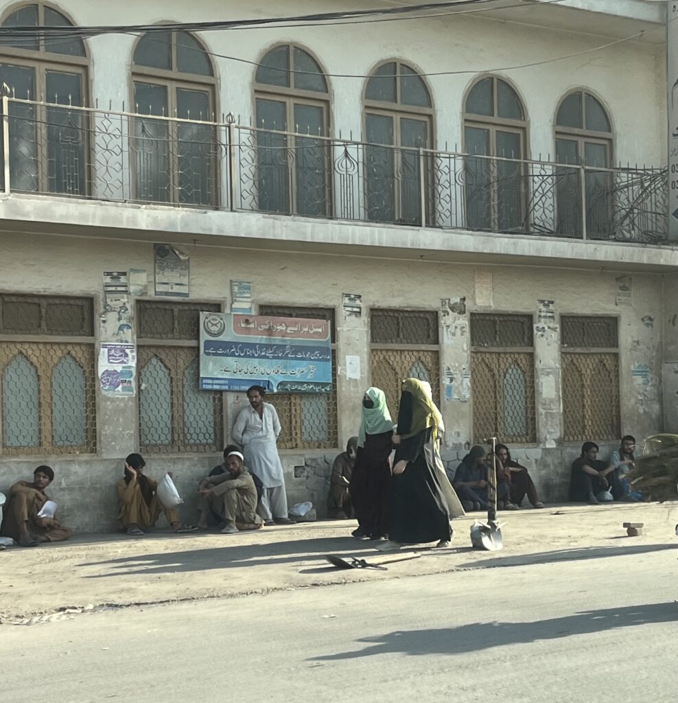 Two women covered from head to toe walk along a street. Both are wearing black abayas and headscarves. One has on a traditional black niqab to cover her face and the other is wearing a black face mask.