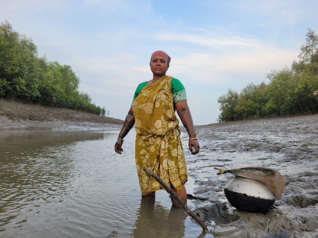 A woman in a yellow sari and green short sleeves stands on the muddy bank of a river submerged up to her knees in water.