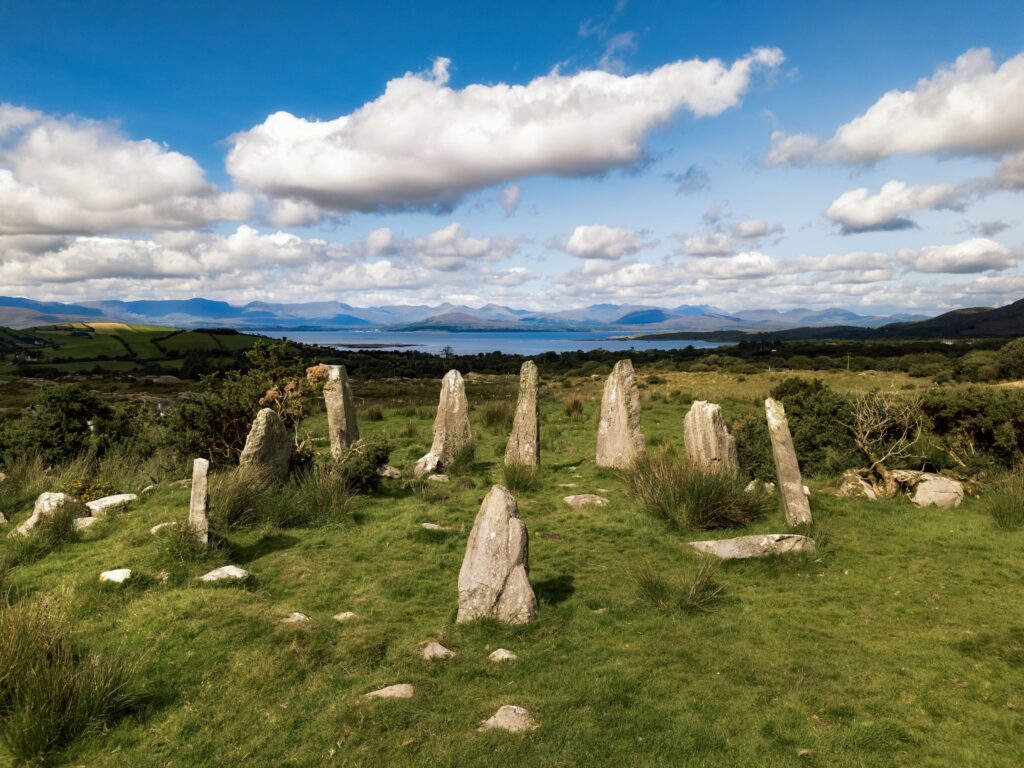 A group of large stones jut out of a wide, green grassy field beneath a vast blue sky with long white clouds floating above.