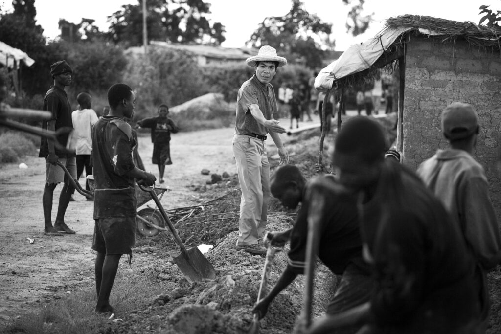 Uma fotografia em preto e branco mostra um homem com chapéu branco, camisa de colarinho e calças cáqui dirigindo o trabalho de um grupo de homens que cavam com pás.