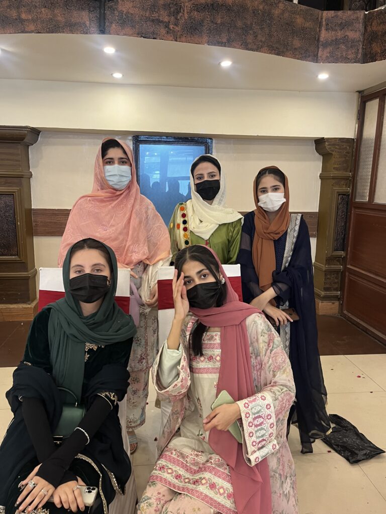 Five women wearing local dresses, headscarves, and face masks pose for a photograph.