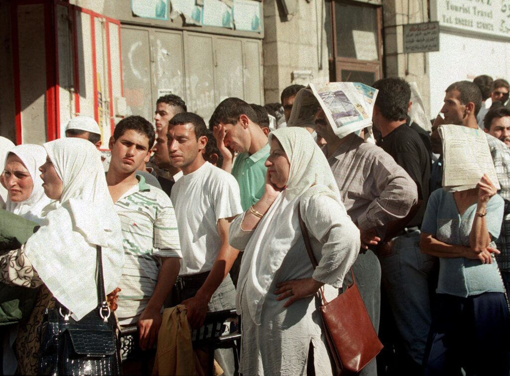 Um grupo de pessoas espera impacientemente do lado de fora de um prédio. As mulheres usam lenços brancos na cabeça, enquanto alguns homens seguram jornais sobre a cabeça para se proteger do sol.