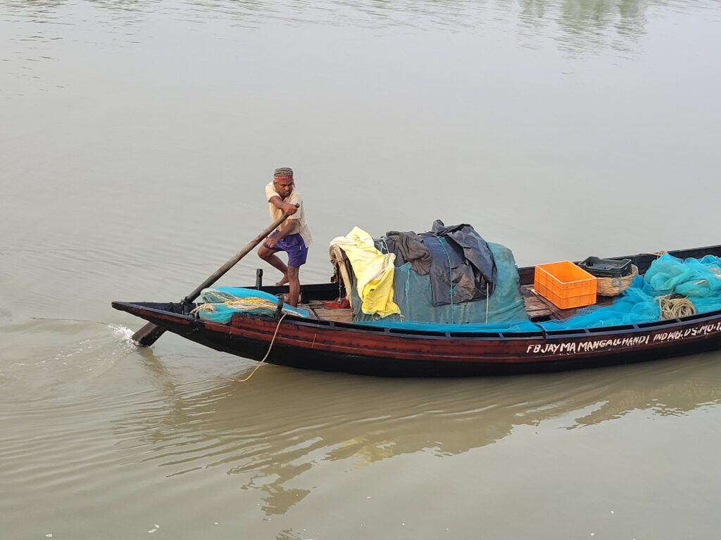A man stands on the bow of a narrow boat laden with fishing gear, steering it with a single oar.