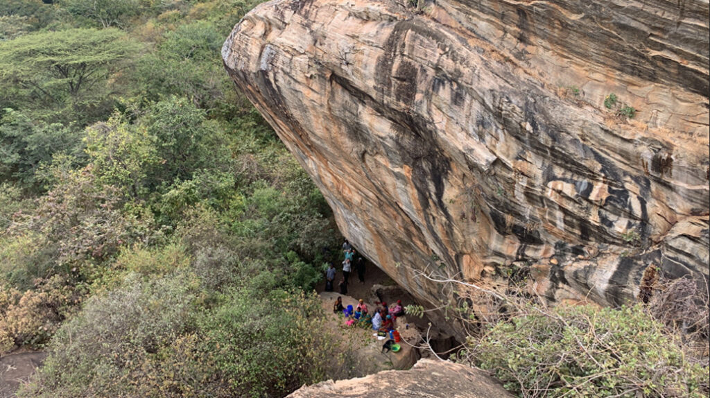 A group of people stand beneath an enormous striped rock towering above them, with lush green fields on the other side.