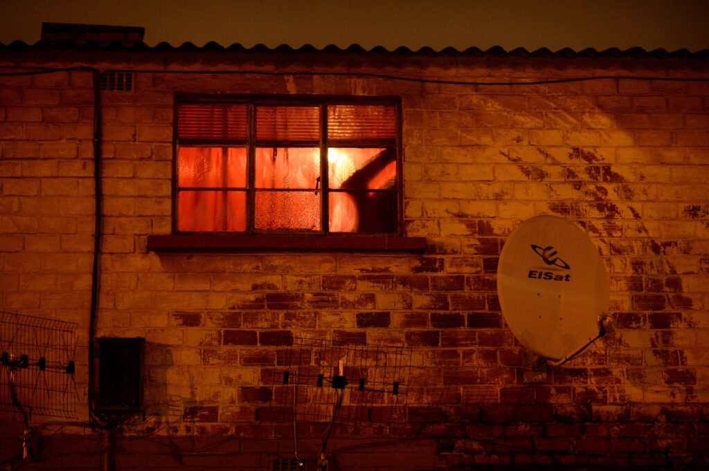 A silhouetted arm can be seen through the window of a brick building bathed in an orangish light.