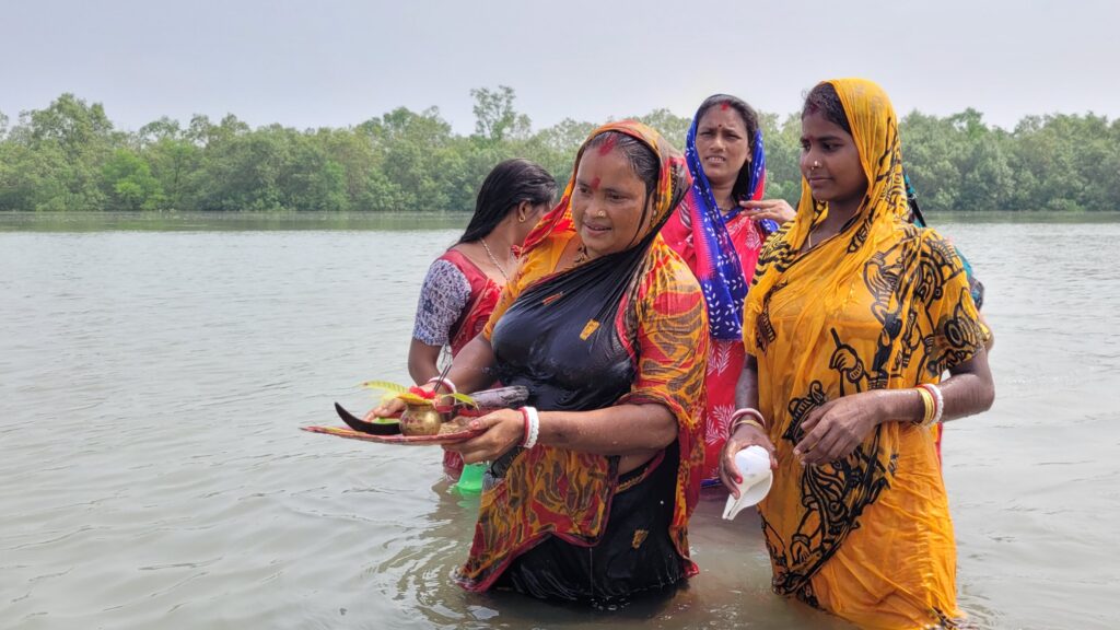 Four women clad in yellow, black, and red saris stand in nearly waist-high water. One of the women holds a tray of small objects.