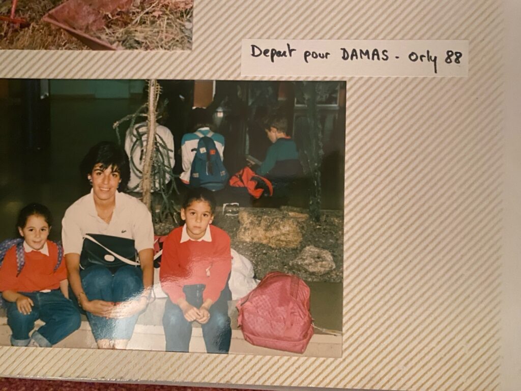 A family photograph in a photo album shows a woman in a white shirt with a purse on her lap sitting next to two young children in red sweaters.
