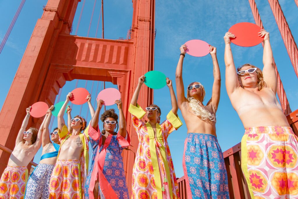 Under a clear blue sky, seven women, several of them topless to reveal their post-mastectomy aesthetic flat closures, stand on an orange-colored suspension bridge holding aloft colorful circles of coral, pink, and green pastel hues. They are clad in loose-fitting colorful clothing with bold patterned prints.