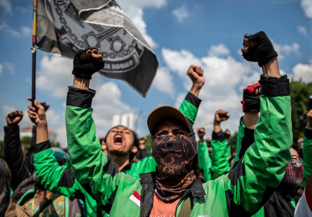 Outside, beneath a blue sky with fluffy white clouds, people wearing bright green jackets hold their fists in the air and wave a flag.