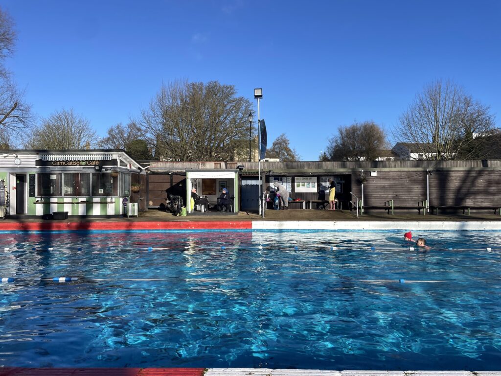 Two people swim in an outdoor pool of shimmering blue water beneath a crisp blue sky. In the background is a row of short structures with outdoor seating and enclosed rooms.