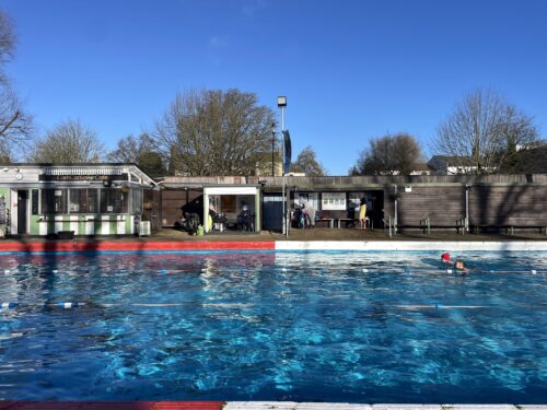 Two people swim in an outdoor pool of shimmering blue water beneath a crisp blue sky. In the background is a row of short structures with outdoor seating and enclosed rooms.